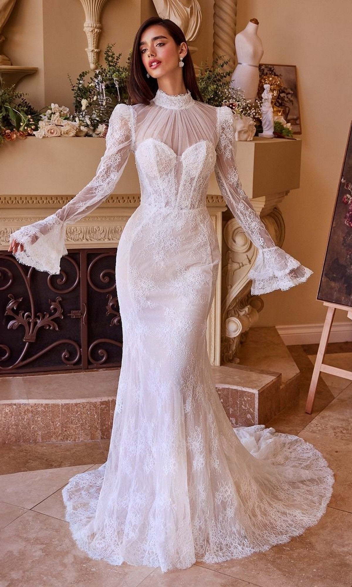 A woman poses indoors by a decorated fireplace wearing the Andrea and Leo WL042 long wedding dress, featuring a strapless lace design, fit-and-flare silhouette, and removable sleeves.